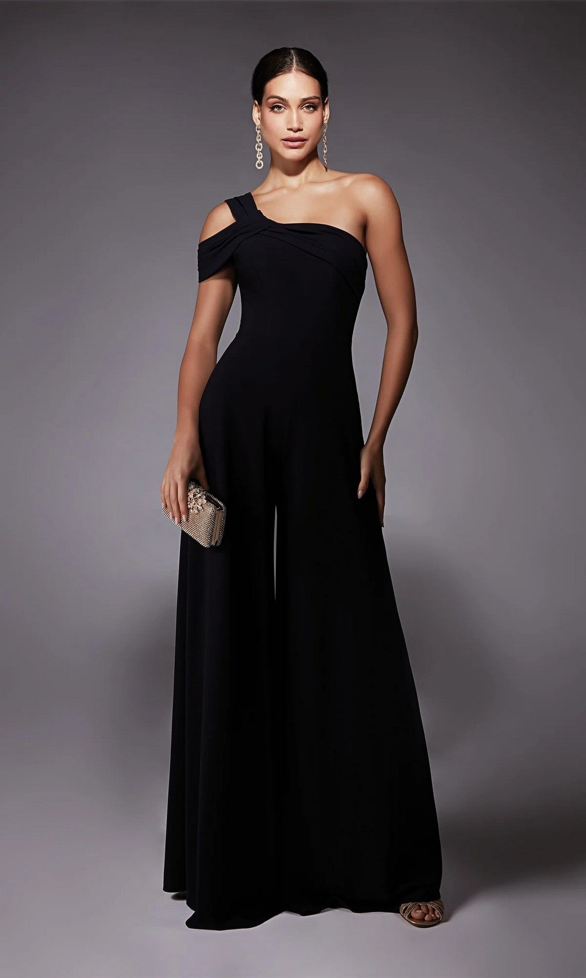 A woman in the Alyce 70066 Long Formal Jumpsuit, featuring a chic one-shoulder design and wide-leg black pants, holds a small clutch against a plain gray background.